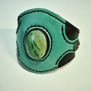 May include: A turquoise leather cuff bracelet with a large oval stone in the center. The stone is green and yellow. The bracelet has black stitching and accents. A silver-colored metal embellishment is on the side.
