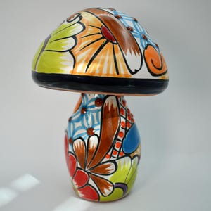 May include: A colorful ceramic mushroom-shaped decorative item. The mushroom cap is painted with orange, white, and green floral designs. The stem features a mix of blue, red, and brown floral patterns. A unique, handcrafted piece.