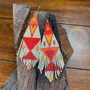 May include: A pair of long, beaded earrings with a geometric pattern in shades of red, orange, and white. The earrings are made with small, seed beads and have a fringe-like design.