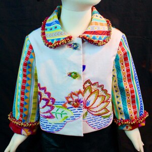 May include: A white jacket with colourful embroidered flowers and geometric patterns on the sleeves and collar. The jacket has a button closure and a fringed trim.