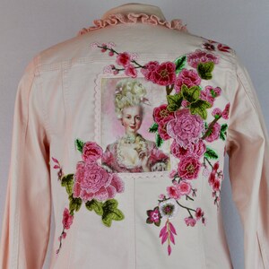 May include: A pink denim jacket featuring embroidered flowers and a vintage portrait of a woman with a large white wig. The jacket has a ruffled collar and a scalloped hem.
