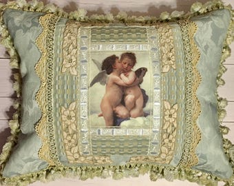 Puede incluir: Una almohada decorativa con tela de damasco verde y blanco. La almohada presenta una imagen enmarcada de dos querubines abrazándose, rodeada de encaje y bordados florales intrincados.