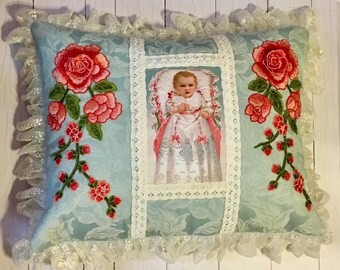 Puede incluir: Una almohada azul claro con ribete de encaje blanco y rosas bordadas. La almohada tiene una imagen enmarcada de un niño con un vestido blanco y una faja rosa. La imagen está rodeada por un borde de encaje blanco.