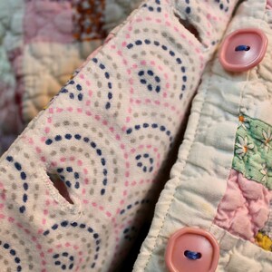 May include: A close-up of a pink and white patchwork quilt with a white fabric flap folded over the top. The flap has a pattern of small pink and blue dots and circles. Two pink plastic buttons are visible on the flap.