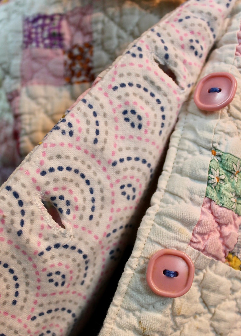 May include: A close-up of a pink and white patchwork quilt with a white fabric flap folded over the top. The flap has a pattern of small pink and blue dots and circles. Two pink plastic buttons are visible on the flap.