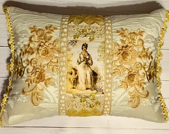 Puede incluir: Cojín decorativo rectangular con un fondo verde claro y bordado floral dorado. El cojín presenta un panel central con una imagen vintage de una mujer, acentuado con encaje y adornos dorados. Los bordes están acabados con encaje amarillo.