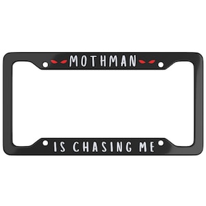May include: Black license plate frame with the text "Mothman is chasing me" in white lettering. Red eyes are depicted on either side of the word "Mothman".