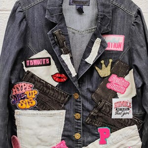 May include: A dark gray denim jacket featuring various patches and fabric appliqués. Patches include text reading IT IS WHAT IT IS, HYPE ME UP SIS, Fashion Junkie, HUMBLE BUT I'M STILL that girl, P, and WHEN GOD MADE ME. Features a gold crown, red lips, and a pink heart.