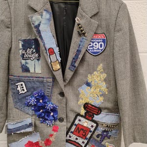 May include: A gray plaid blazer with denim patches and decorative embellishments. Patches include a lipstick, a perfume bottle, a star, and the text "a lil' bougie". The blazer also features a blue flower arrangement and an Interstate 290 sign.