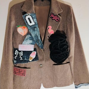 May include: A brown blazer with various patches, including a peach, a flamingo, and the word "Hustler." The blazer has a black flower detail and denim accents. The text "BEHIND EVERY SUCCESSFUL WOMAN IS HERSELF" is also visible.
