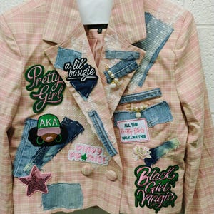May include: A light pink plaid blazer adorned with various patches and denim fabric scraps. Patches include text reading Pretty Girl, a lil bougie, AKA, ALL THE Pretty Girls WALK LIKE THIS, PINKY PROMISE, and Black Girl Magic, alongside a sequined star.