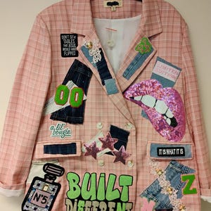 May include: A pink plaid blazer featuring various patches including a sequined pink lip, a perfume bottle, and text reading "BUILT DIFFERENT," "DON'T SIT AT TABLES THAT JESUS WOULD HAVE FLIPPED," "LIMITED EDITION," and "IT IS WHAT IT IS" on the front.