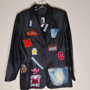 May include: Black blazer adorned with patches and denim accents. Patches include text that reads "LOVE The Skin You're In", "BLACK Beautiful BLESSED", and a large "M". Features a denim pocket and a perfume bottle patch.