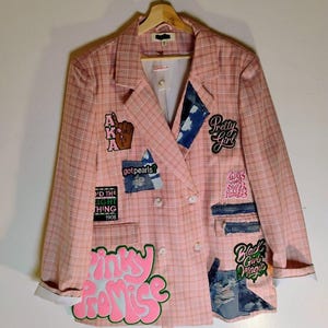 May include: Pink plaid blazer with multiple patches. Patches include text that reads "AKA", "Pretty Girl", "Pinky Promise", "Girls Super Powers", "Black Girl Magic", and "'D THE LIGHT THING 1908". The blazer has a double-breasted design.