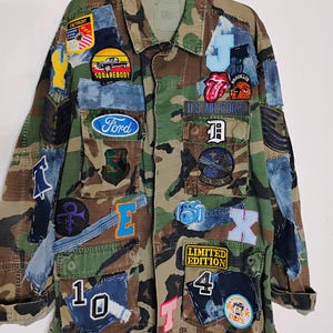 May include: A camouflage print jacket featuring numerous colorful patches, including a Ford logo, a Detroit emblem, a Squarebody truck graphic, a Rolling Stones tongue, a camera, and various letters and numbers like 10, 4, J, E, T, and X.
