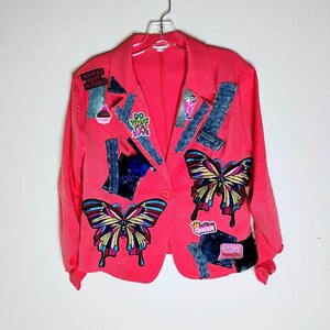 May include: A bright pink blazer adorned with colorful butterfly patches and denim accents. The jacket features various embroidered patches with text, including "Do What You Love" and "Booked, Busy, Blessed."