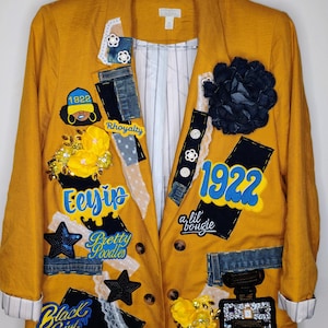 May include: A mustard yellow blazer featuring various appliqués, including denim patches, yellow fabric flowers, a dark blue fabric flower, and embroidered text such as 1922, Eeyip, Pretty Poodles, a lil' bougie, and Black Girl Magic, with a white pinstriped lining.