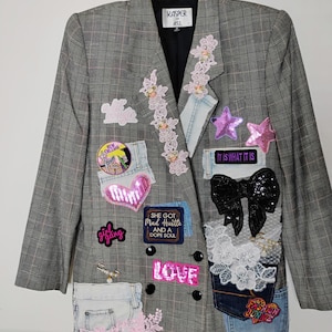 May include: A grey plaid blazer featuring various decorative patches, including pink sequins, denim pocket accents, lace floral appliqués, a black sequined bow, and text patches reading "IT IS WHAT IT IS," "SHE GOT MAD HUSTLE AND A DOPE SOUL," and "LOVE."