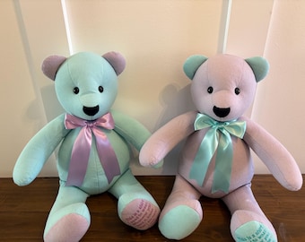 Könnte beinhalten: Zwei Plüschteddys. Einer ist hellblau mit Lavendelakzenten und einer Lavendelschleife. Der andere ist Lavendel mit hellblauen Akzenten und einer hellblauen Schleife. Beide haben schwarze Augen und gestickte Smileys. Die Bären sitzen auf einer Holzoberfläche.