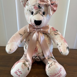 May include: A floral print teddy bear with a pink bow on its head and a matching bow around its neck. The bear's paws are embroidered with the name "Isla Rose" and the date "October 15th, 2024". The bear is made of soft fabric.