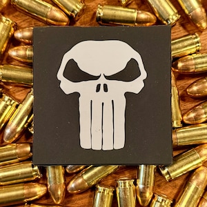 May include: A black square patch with a white skull design, encircled by numerous brass-coloured bullets. The skull has large eye sockets and vertical lines for teeth. The bullets are arranged on a wooden surface.