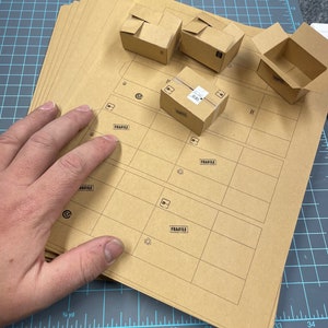 May include: A stack of brown cardboard sheets with printed guidelines for assembling boxes. There are four small brown cardboard boxes with the word "Fragile" printed on them. The boxes are on a blue and white grid surface.