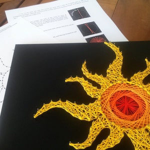 Op de afbeelding: Een string art zon met gele, oranje en rode draad op een zwarte achtergrond. De zon heeft een cirkelvormig midden en acht stralen.