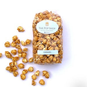May include: A clear plastic bag filled with caramel popcorn. The bag has a white label with the text "THE POP SHOP" and the words "Life's short, eat good popcorn". The label also has a small cupcake icon. The bag is sitting on a white surface. There is a small amount of popcorn scattered around the bag.