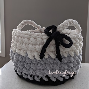 May include: A white, gray, and black crocheted basket with a black bow. The basket has two handles and is made with a chunky yarn. The text "LoveDIncyDesigns" is visible on the bottom of the basket.