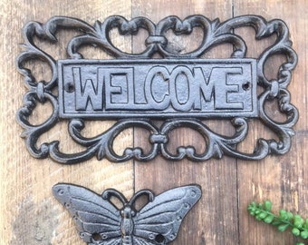 Puede incluir: Un letrero de bienvenida de hierro fundido negro con adornos de volutas y un gancho en forma de mariposa. El letrero dice "WELCOME" en letras en relieve.