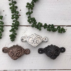 Könnte beinhalten: Drei verzierte Schubladenknöpfe aus Gusseisen in verschiedenen Ausführungen: Schwarz, Weiß und Braun. Die Knöpfe haben ein dekoratives, rollenförmiges Design.