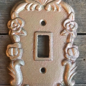 May include: A gold-colored metal light switch cover with a decorative floral design. The cover features a raised design of roses and leaves.