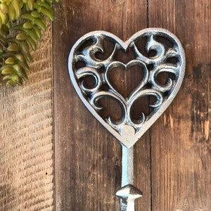 May include: A silver metal heart-shaped hook with a decorative scroll design. The hook is mounted on a wooden surface.