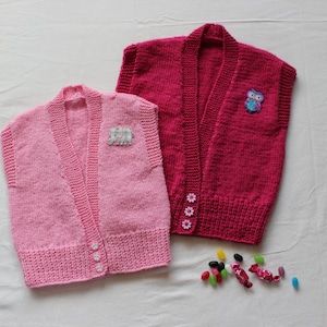 May include: Two hand-knitted children's vests. One is light pink with three white buttons and a small patch. The other is a darker pink with an owl patch and flower buttons. Both have a V-neck design.