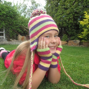 May include: A young girl wearing a striped knit hat with earflaps and matching fingerless gloves. The hat and gloves are pink, yellow, green, and purple. The girl is lying on her stomach in a grassy area and smiling.