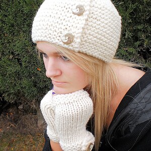 Puede incluir: Un gorro de punto blanco con dos botones plateados. El gorro lo lleva puesto una persona con el pelo largo y rubio. La persona lleva un jersey negro.