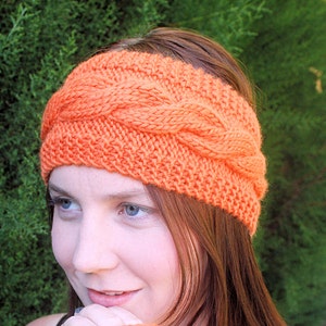 May include: A bright orange knitted headband with a cable knit pattern. The headband is worn by a person with long, brown hair.