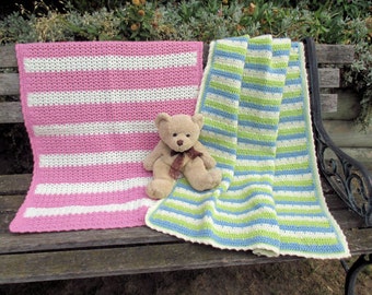 May include: Two crocheted baby blankets, one pink and white striped and one blue, green, and white striped, are displayed on a wooden bench with a brown teddy bear sitting on top of the blue blanket.