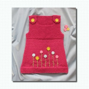 Op de afbeelding: Een gebreide roze jurk met gele en witte bloemversieringen. De jurk heeft een vierkante halslijn, schouderbandjes met gele bloemknoopjes en geborduurde bloemstelen. Extra bloemknoopjes zijn bij de jurk geplaatst.