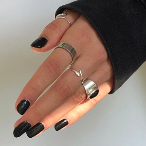 May include: A hand with black nail polish is wearing five silver rings. The rings are different styles, including a thick band, a thin band, a ring with a pointed design, and a ring with a stone.