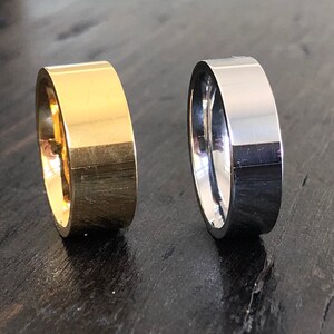 May include: Two simple wedding bands, one gold and one silver, on a dark wood surface.