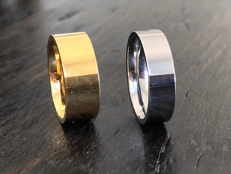 May include: Two simple wedding bands, one gold and one silver, on a dark wood surface.