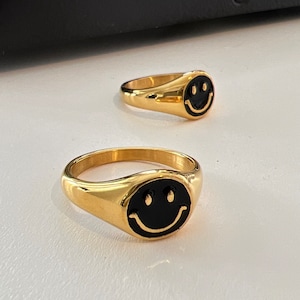 May include: Two gold signet rings with black enamel smiley faces. The rings are on a white surface.