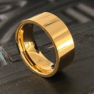 May include: A gold-colored metal ring with a flat, wide band. The ring is smooth and shiny.