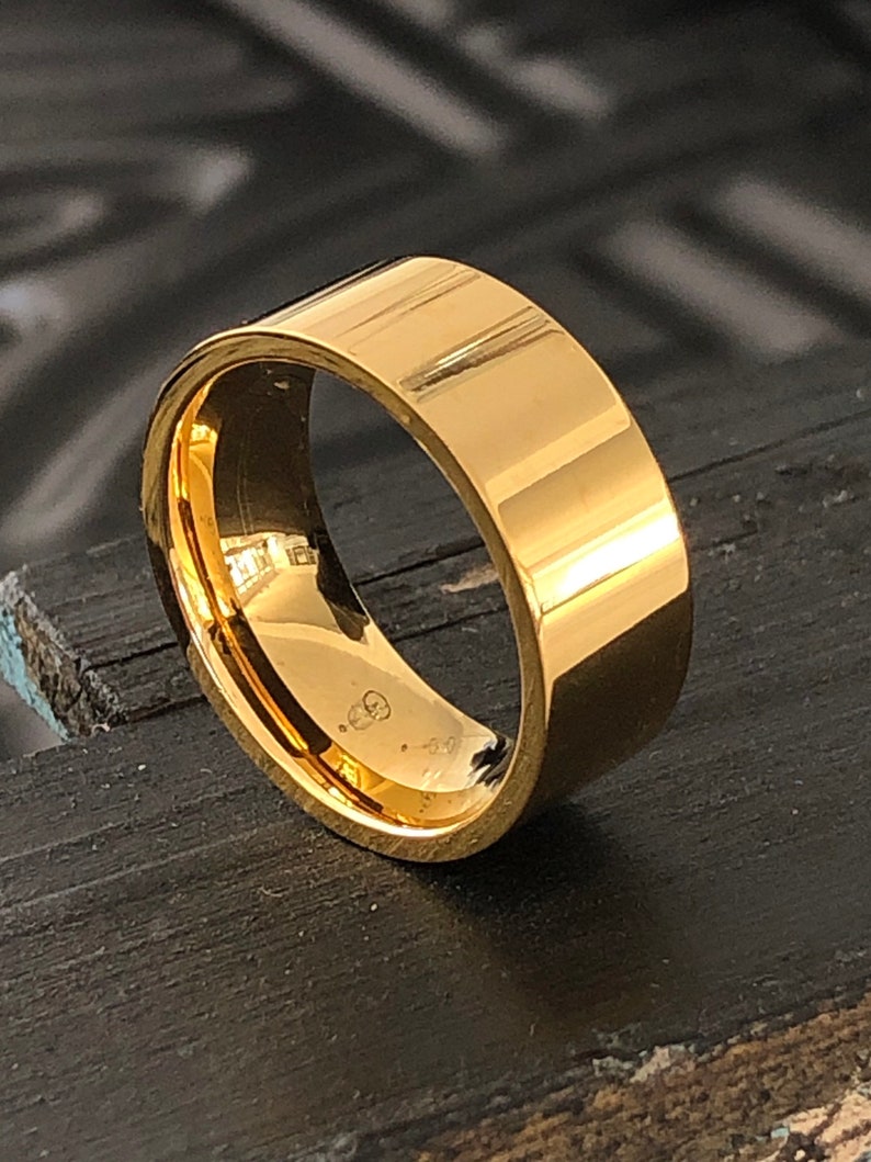 May include: A gold-colored metal ring with a flat, wide band. The ring is smooth and shiny.