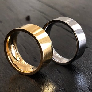 May include: Two wedding bands, one gold and one silver, are shown on a dark wooden surface. The bands are both simple and classic in design.