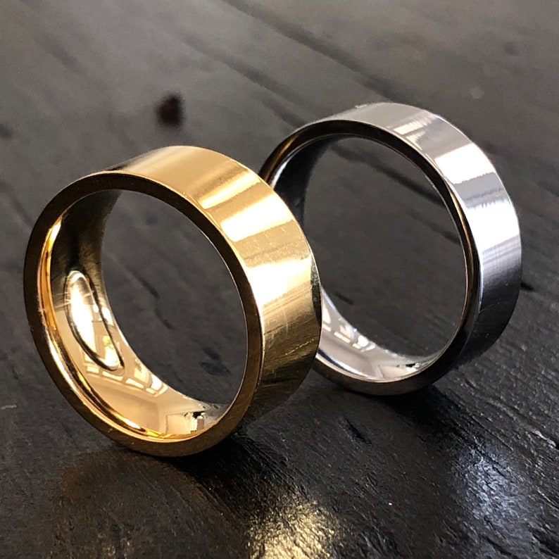 May include: Two wedding bands, one gold and one silver, are shown on a dark wooden surface. The bands are both simple and classic in design.