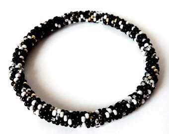 May include: A circular beaded bracelet constructed from a dense arrangement of small, round beads in a repeating pattern of matte black, glossy white, metallic silver, and metallic gold, creating a textured, continuous loop design.