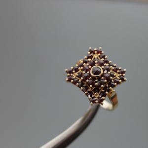 Può includere: Un anello d'oro con una montatura quadrata che presenta un gruppo di pietre di granato rosso.