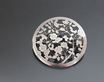 May include: A silver brooch with a delicate floral design. The brooch is circular and features a stylized tree with leaves and flowers.